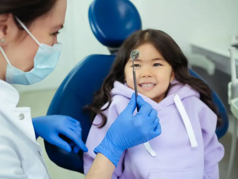 Smiling girl in a dental chair while a hygienist holds up a small dental mirror
