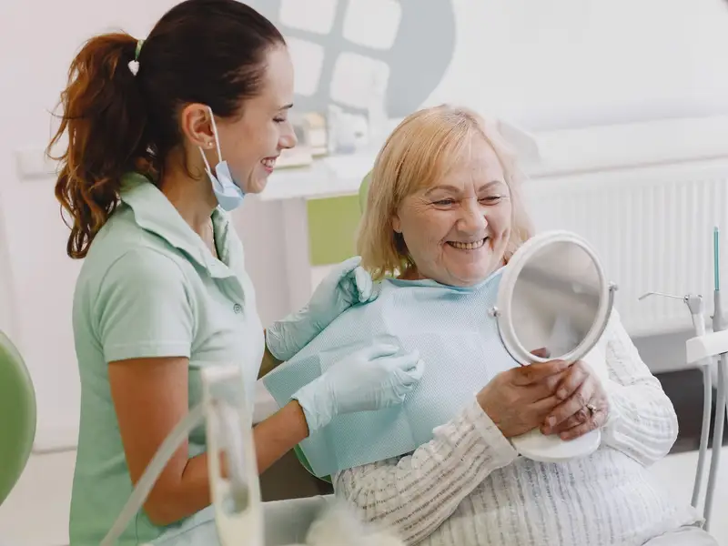 Older woman smiling into a hand mirror beside a dental hygienist