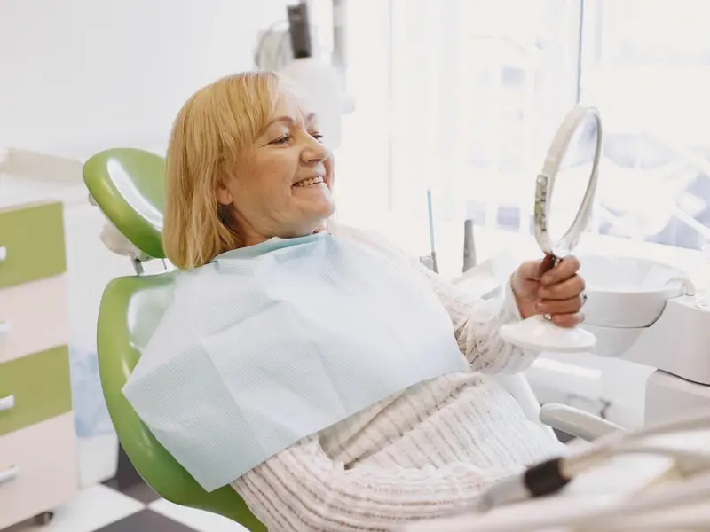 Older woman in a dental chair smiling at her teeth in a hand mirror