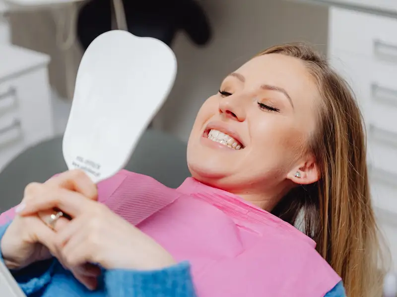 Woman in a dental chair laughing as she looks at her smile in a hand mirror
